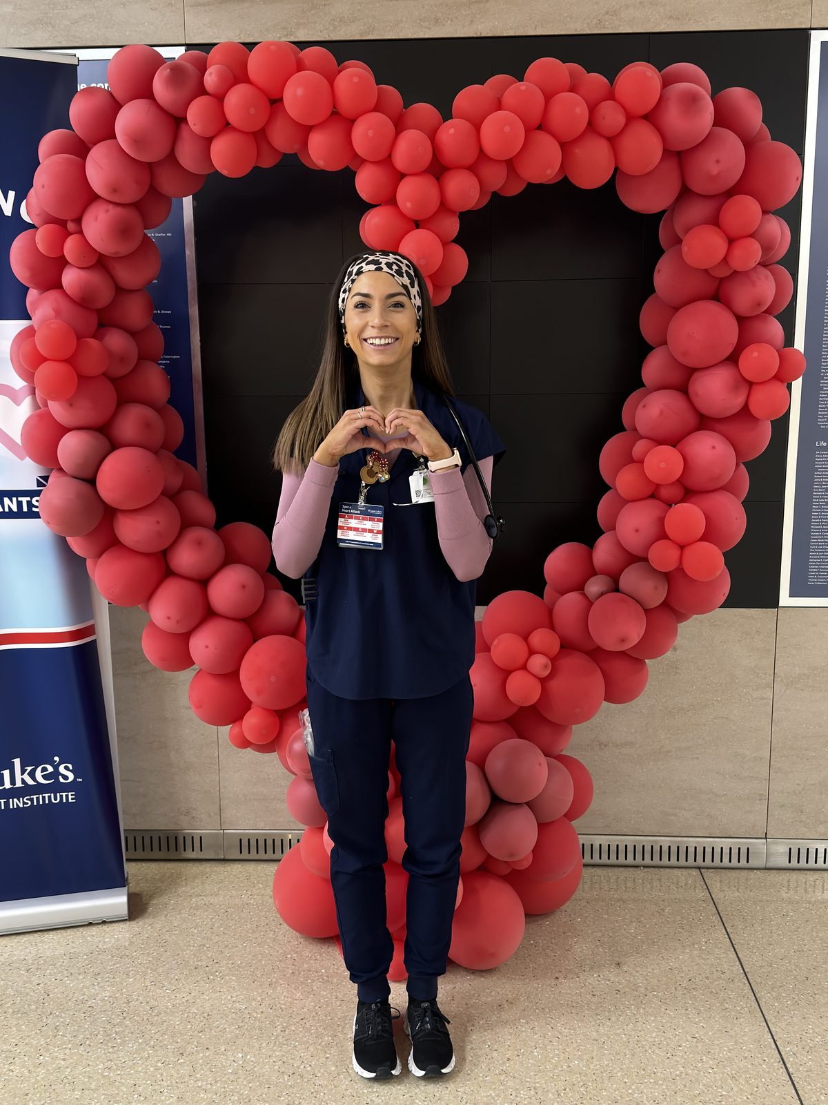 Josie in scrubs making a heart with her hands inside a red balloon heart